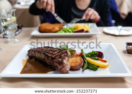 Delicious strip loin beef steak meal served with fresh vegetables and marsala portobello mushroom sauce. Focus on steak and deliberate shallow depth of field on subject having a meal across the table.