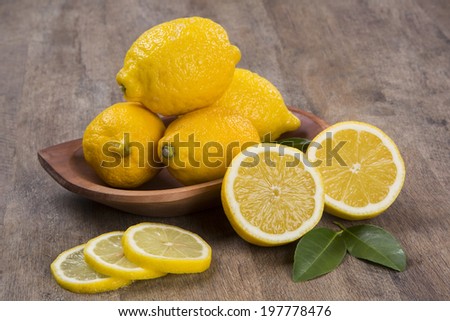 A sliced lemon and a lemon cut in a half in front of a pot of lemons over a wooden surface.