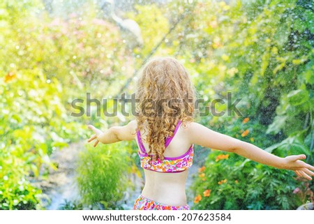 Little girl under water splashes in the summer garden. View from the back.