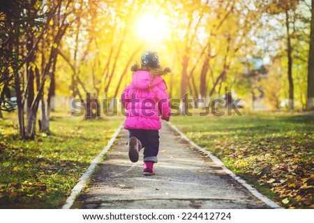 Little girl in a red jacket, running away the road in the autumn park.