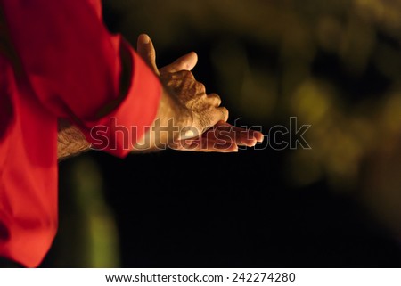 Close up of the clasped hands of a male flamenco dancer wearing a red shirt in the darkness which could indicate clapping, anxiety or washing them, view from behind