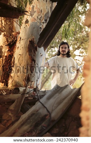 Long Hair Young Man in Casual White Shirt Behind Big Tree, Looking at the Camera  Behind the Tree