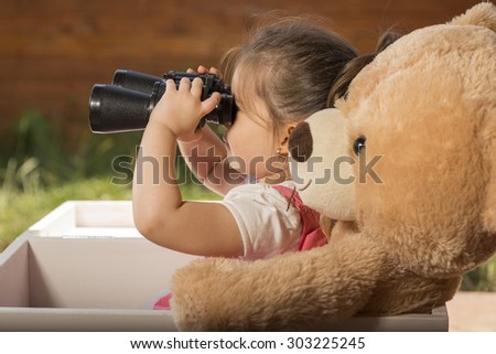 Beautiful little girl with binoculars inside white wooden box with toy bear exploring surroundings pretending to be on safari
