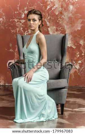 Young beautiful brown-haired woman in blue light silk dress with open shoulders and a long skirt and sandals sitting in the chair