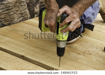 A construction workerÃ¢Â?Â?s hands using a power drill to secure wood boards to a patio deck.