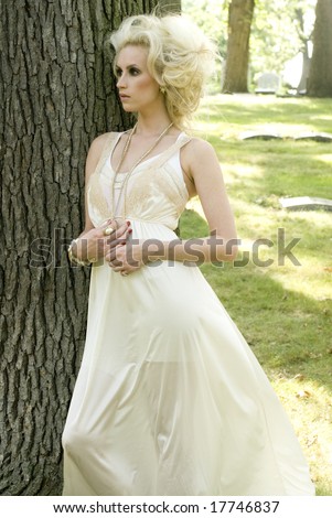 Tall blonde woman with semi-sheer white dress standing against a tree