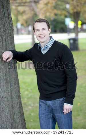 An attractive young male model posing by a tree wearing a sweater and a button down.