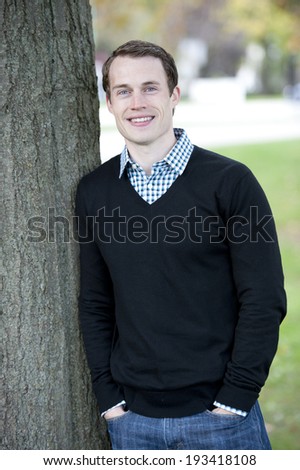 An attractive young male model posing by a tree wearing a sweater and a button down.