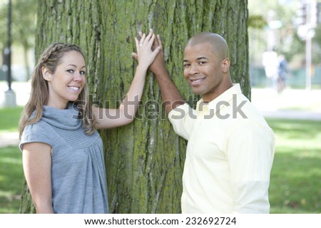 A happy young couple posing on a sunny day next to a large green tree.