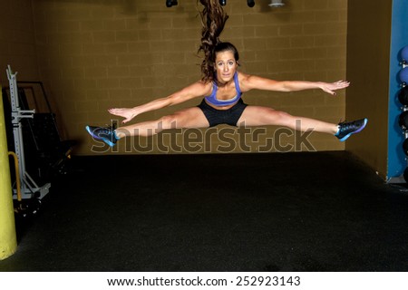 An beautiful athletic and toned brunette doing a jump split in a gym.