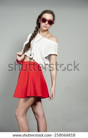 Attractive young brunette girl wearing a pair of red sunglasses, white tank top and a red mini skirt on a gray studio background.