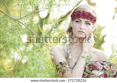 A portrait of a young gorgeous blonde girl looking up with her hand on her cheek while wearing a red flower crown with a flowery top with the sun shining behind her.
