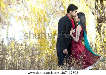A young Indian man kissing the forehead of his Indian bride who is wearing a Sari and both are standing under a willow tree.