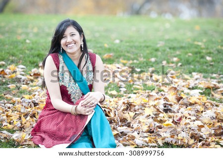A young happy Indian girl sitting in a pile of leaves outdoors on a cloudy day.