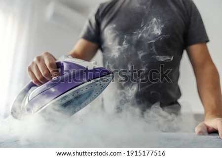 The hot steam from the iron. Powerful film effect of steam on photography. A close-up of a man's body in a grey t-shirt ironing clothes on an ironing board
