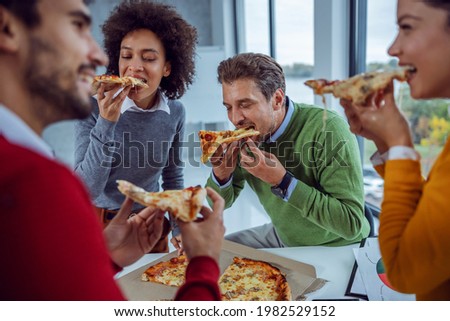 Pizza time. An excited multicultural group of businesspeople standing in the office eating pizza for lunch. Team building during office break. They are eating pizza, enjoying pizza