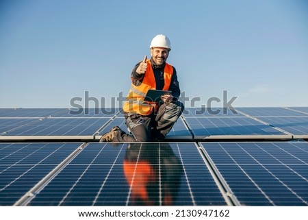 A worker on rooftop kneeling next to solar panels with tablet in hands and giving thumb up for sustainable life. Stock fotó © 