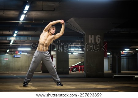 Handsome muscular young man stretching at parking garage, natural lights, dark place.