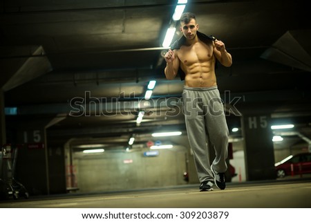 Shirtless handsome muscular young man walking at parking garage, natural lights, dark place.