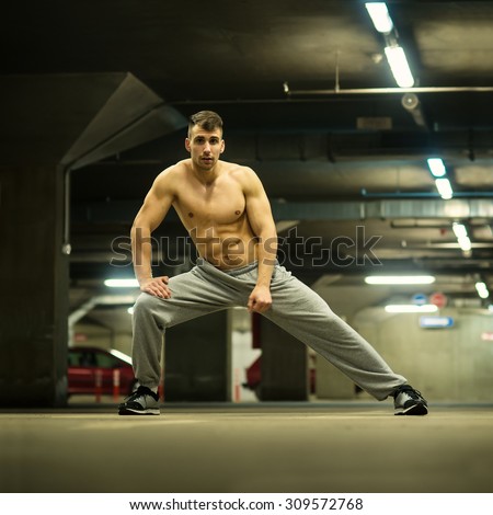 Shirtless handsome muscular young man stretching at parking garage, natural lights, dark place. Shallow depth of field.