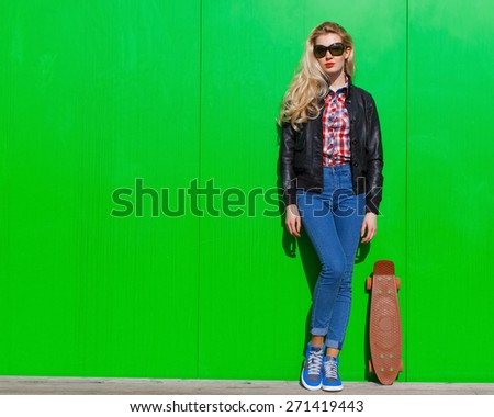Beautiful blonde girl in huge sunglasses and a black jacket posing next to green wall on a sunny day with a small skateboard
