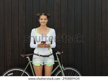 Pretty girl in shorts and t-shirt stands with coffee cup and bicycle fix gear near the wall of wooden planks bright sunny day. Outdoor. Warm colors