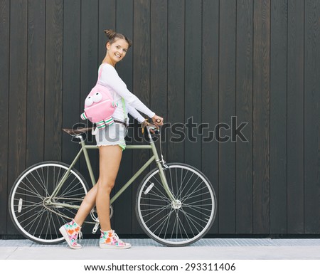 Pretty girl in shorts and t-shirt with a funny pink backpack stands with his bicycle fix gear nex to the wall of wooden planks sunny day