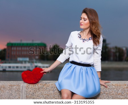 Beautiful fashionable Girl with red heart in the park in warm summer evening sitting on a stone bank of the river alone with a red heart
