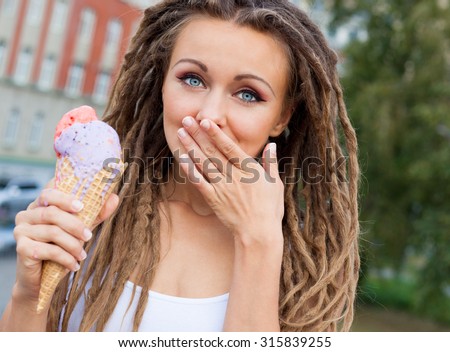 Beautiful girl with dreadlocks eating colorful ice cream and covers her mouth a palm on a warm summer night in the street. Outdoor