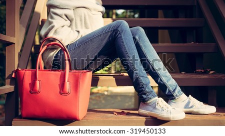 Girl sitting on the stairs with a big red super fashionable handbags in a sweater jeans and sneakers on a warm summer evening. Warm colors