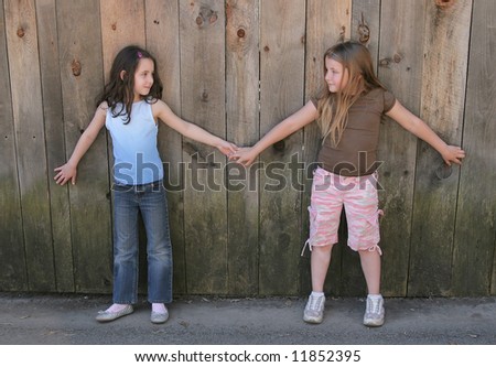 two young girls holding hands and playing against a wooden fence