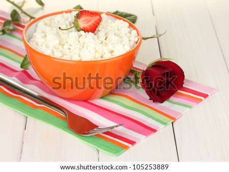 cottage cheese with strawberry in orange bowl, fork and flower on colorful napkin on white wooden table close-up
