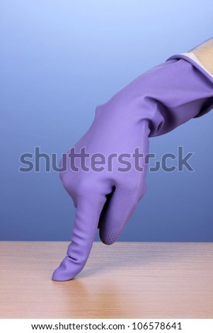 Hand in color cleaning glove holding finger over surface of wooden table on blue background