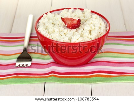 cottage cheese with strawberry in red bowl and fork on colorful napkin on white wooden table close-up