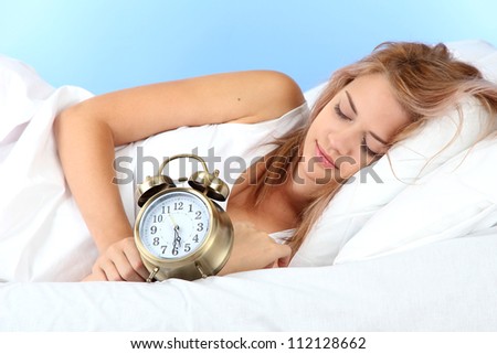 young beautiful woman sleeping on bed with alarm clock on blue background
