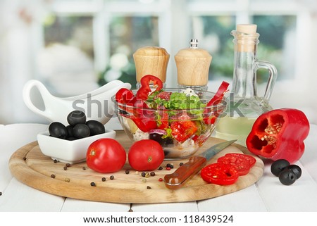 Fresh greek salad in glass bowl surrounded by ingredients for cooking on wooden table on window background