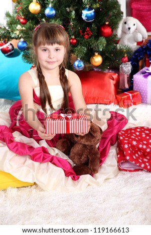 Beautiful little girl in holiday dress with gift in hands in festively decorated room