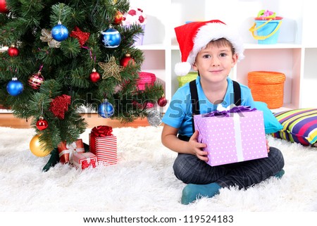 Child in Santa hat sits near Christmas tree with gift in hands