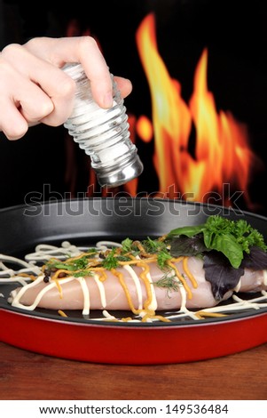 Hand adding spices to raw chicken fillets on dripping pan, on fire background