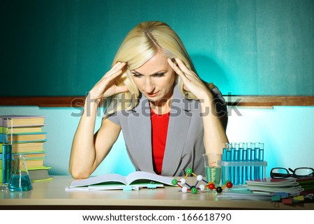 Tired chemistry teacher sitting at table on blackboard background
