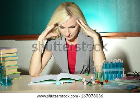 Tired chemistry teacher sitting at table on blackboard background