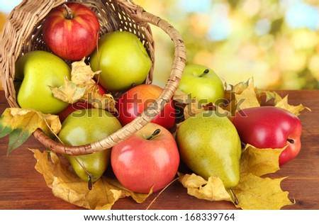 Beautiful ripe apples and pears with yellow leaves in basket on table on bright background
