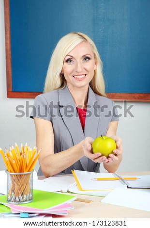 School teacher with apple sitting at table on blackboard background