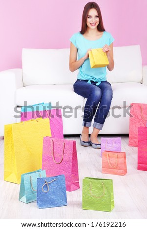 Beautiful young woman sitting on sofa with shopping bags on pink background