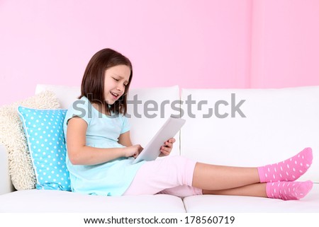 Beautiful little girl sitting on sofa with tablet, on home interior background