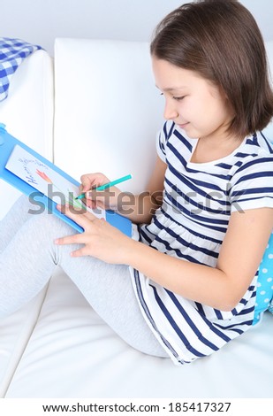 Beautiful little girl sitting on sofa and drawing picture, on home interior background