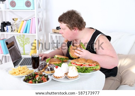 Fat man has a big lunch and playing games on laptop, on home interior background