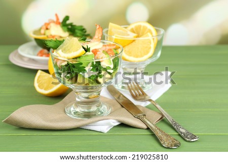 Tasty salads with shrimps and avocado in glass bowl and on plate, on wooden table, on bright background