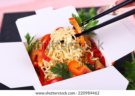 Chinese noodles and sticks in takeaway box on black mat on pink background