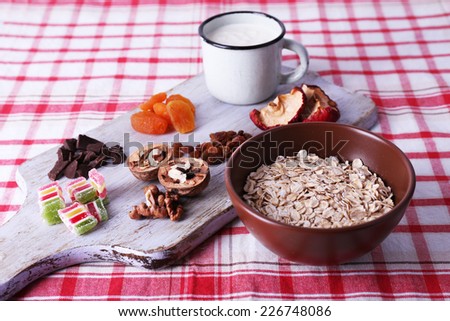 Bowl of oatmeal, mug of yogurt, marmalade, chocolate, raisins, dried apricots and walnuts on wooden cutting board on checkered fabric background